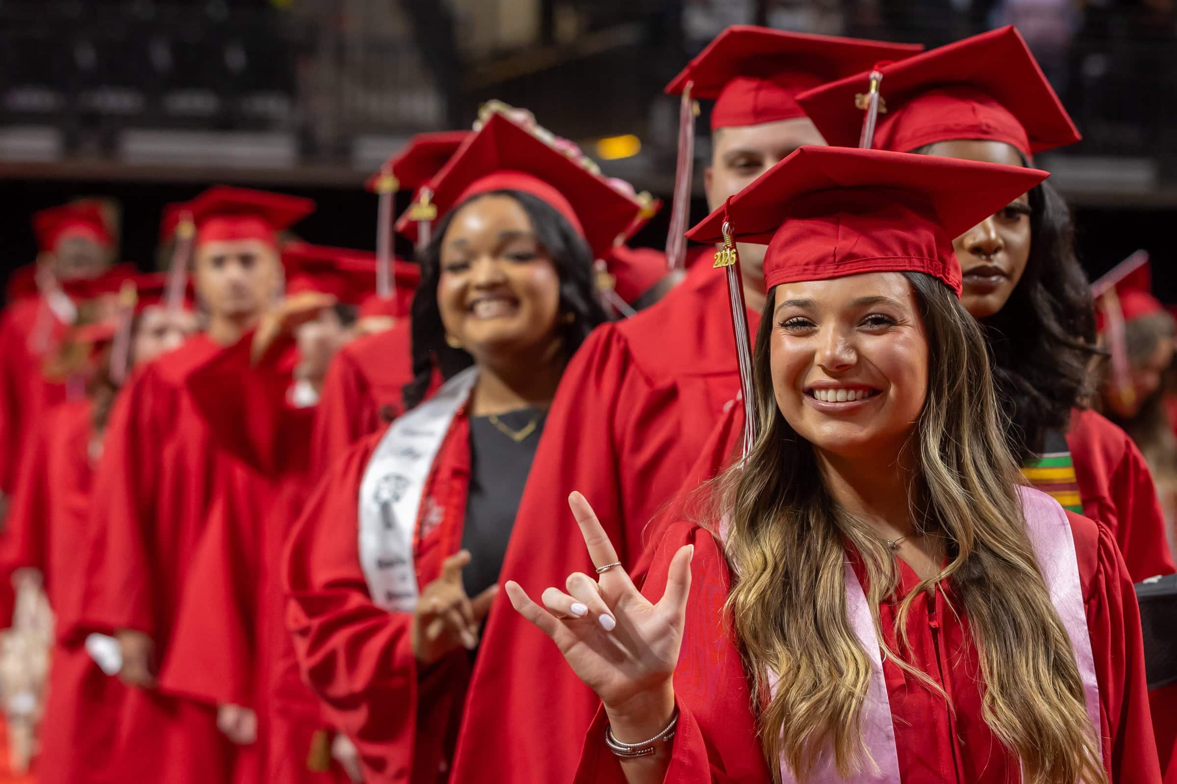 Smiling graduates in red caps and gowns celebrate commencement; a woman in front flashes a hand sign as classmates gather behind her.
